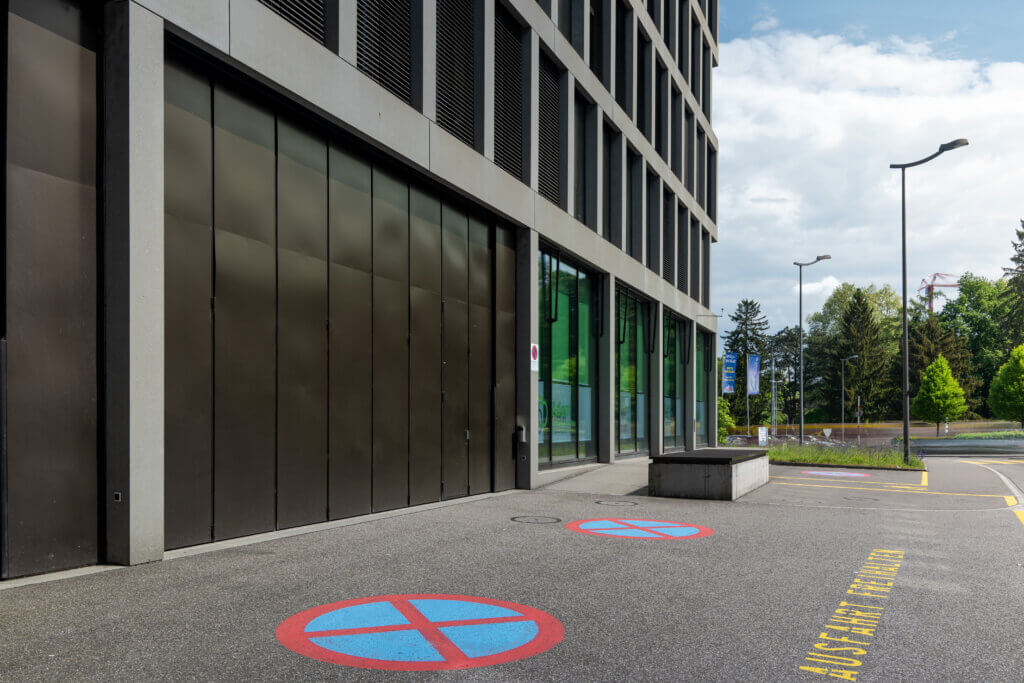 Urbane Gebäudefassade mit großen Fenstern und modernem Design. Auf dem Gehweg in der Nähe des Eingangs sind zwei Parkverbotssymbole aufgemalt. Im Hintergrund sind Bäume und Straßenlaternen zu sehen, was es zu einer perfekten Eventlocation für jeden Firmenanlass macht, der im CAMPUSSAAL stattfindet.