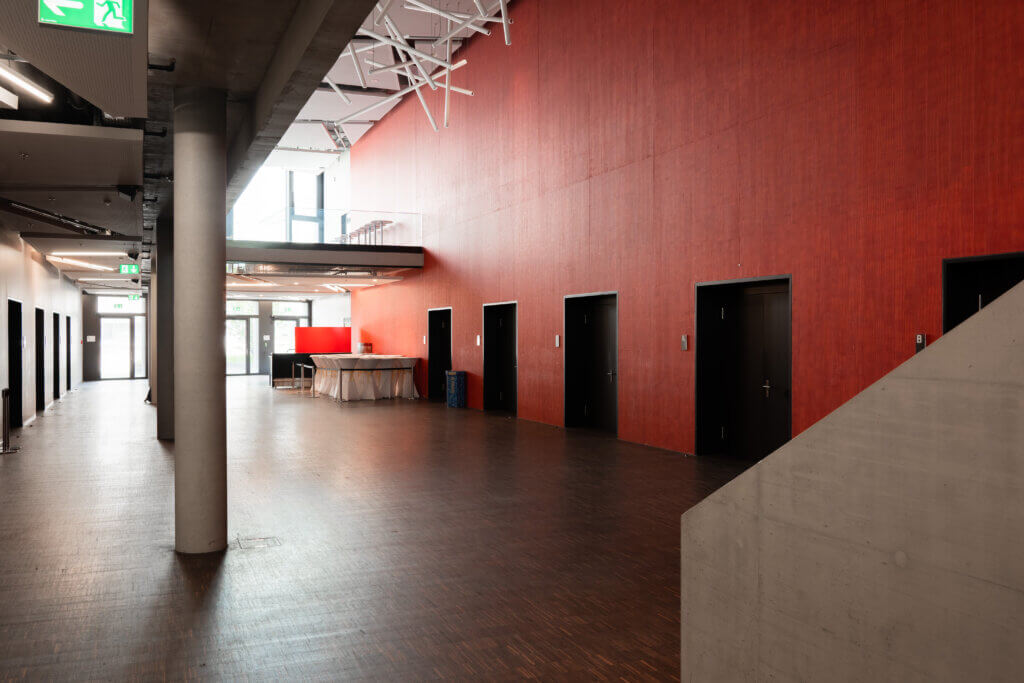 Ein geräumiger, moderner Innenflur mit hoher Decke, roten Wänden, mehreren schwarzen Türen und Betonboden gibt den Ton für diese erstklassige Eventlocation an. Ein Ausgangsschild und eine weiße Treppe sind sichtbar und machen sie zur idealen Wahl für Ihren nächsten Firmenanlass bei CAMPSUSAAL.