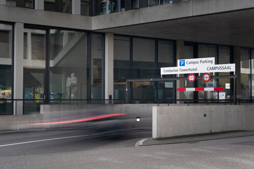 Eine Straßenansicht, die ein Betongebäude mit Hinweisschildern zum Campus Parking, zum Centurion Towerhotel und zum CAMPUSSAAL, der beliebten Eventlocation, zeigt. Auf der Straße bewegt sich ein verschwommenes Fahrzeug.