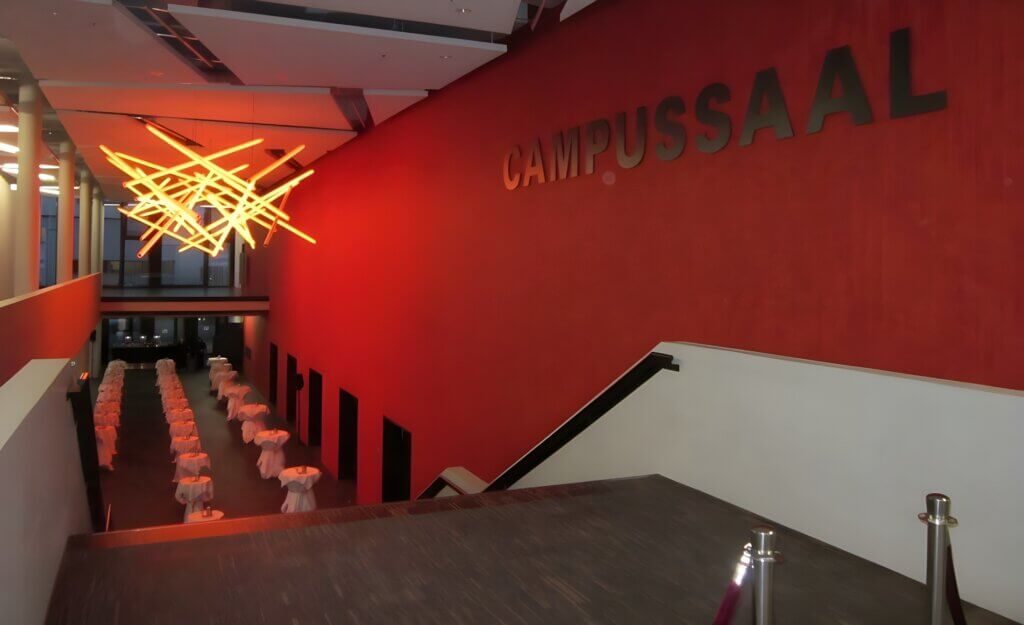 Ein schwach beleuchteter Flur mit roten Wänden, einer hellen Lampe und an beiden Seiten aufgereihten Tischen mit weißen Tischdecken führt zu einem „CAMPUSSAAL“-Schild und macht ihn zur perfekten Hochzeitslocation.