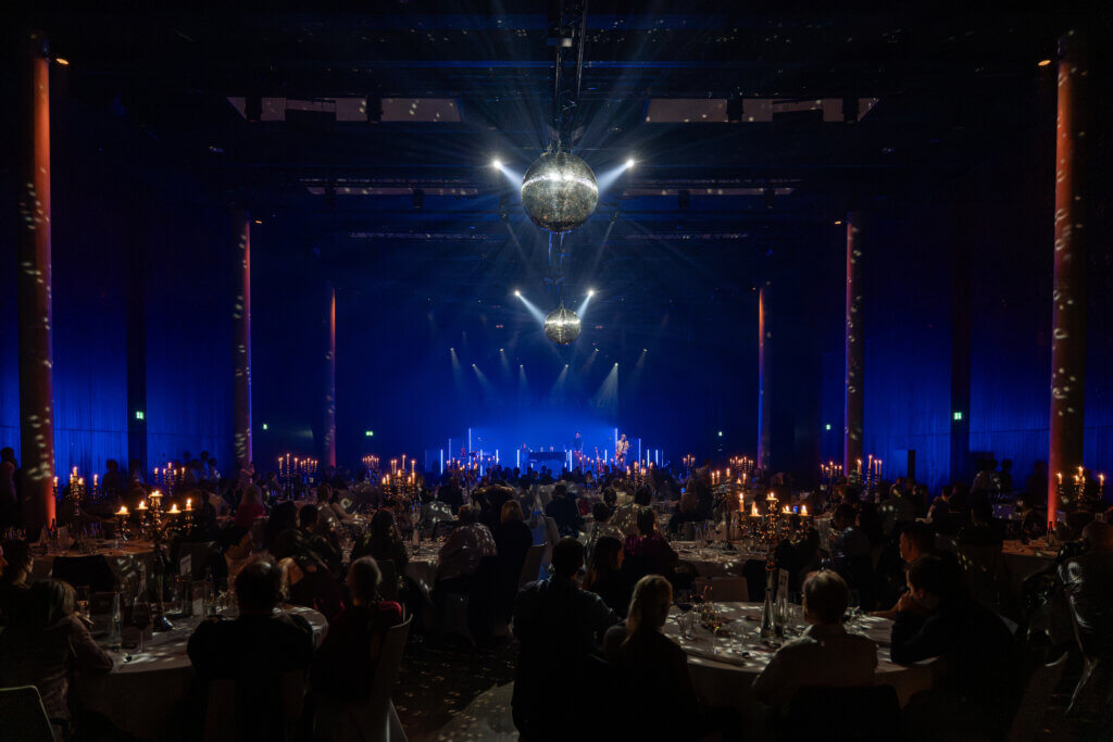 Ein schwach beleuchteter Bankettsaal mit Gästen an runden Tischen, Kerzen auf den Tischen und Discokugeln, die das Licht über einer Bühne mit blauer Beleuchtung reflektieren.