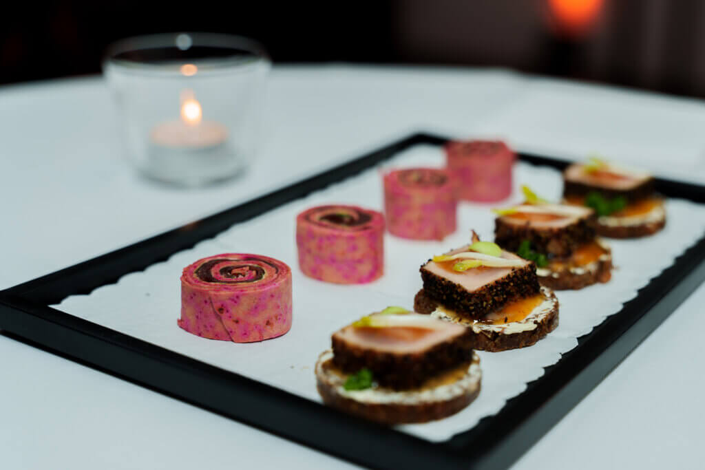Apéro für 100 bis 500 Personen im Aargau: Locations, Kosten und Planung 2 Ein Tablett mit verschiedenen mundgerechten Häppchen, darunter gerollte rosa Strudel und geschichtete Stücke auf rundem Brot, präsentiert auf einem schwarzen Tablett mit einer brennenden Kerze im Hintergrund.
