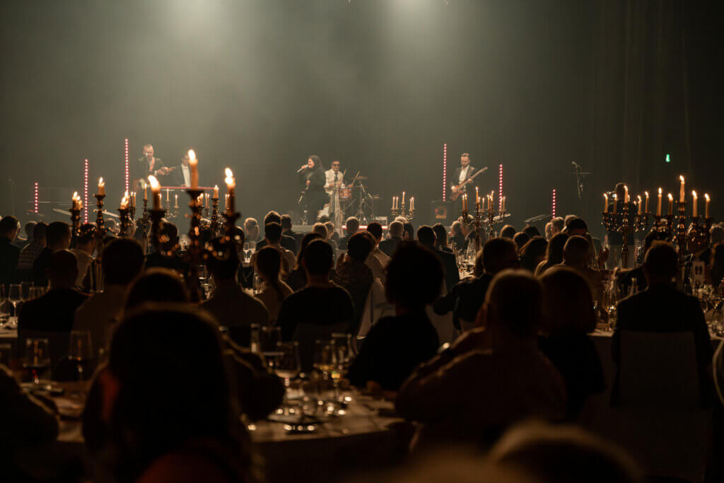 An runden Tischen mit Kerzen sitzendes Publikum, das in einem schwach beleuchteten Saal eine Live-Musikdarbietung auf der Bühne verfolgt.