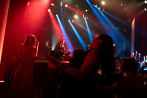 Menschen auf einer schwach beleuchteten Veranstaltung mit Bühnenlicht und einer Band im Hintergrund, während die Teilnehmer Kontakte knüpfen und die Atmosphäre genießen.