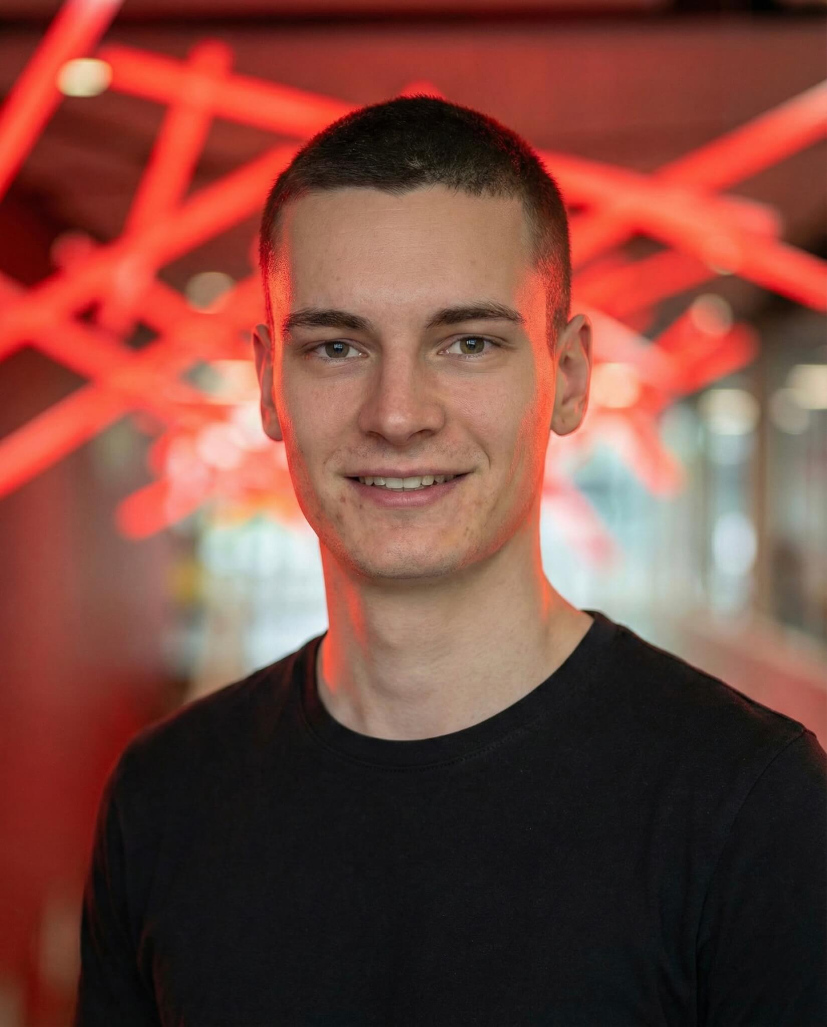 Ein junger Mann mit kurzen dunklen Haaren und einem schwarzen T-Shirt lächelt in die Kamera. Er steht in einem Innenraum mit roten Neonröhren im Hintergrund, die eine moderne Unternehmensatmosphäre verbreiten.